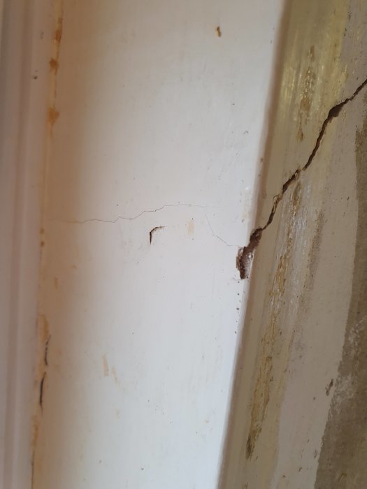 Sprickor i en nermald vägg vid ett hörn som avslöjar underliggande skikt, tecken på möjliga byggproblem.