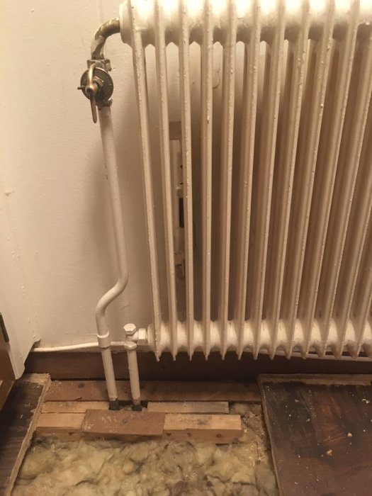 Ett upprivet golv i ett kök med synliga elementrör och isolering, samt en radiator ovanför.