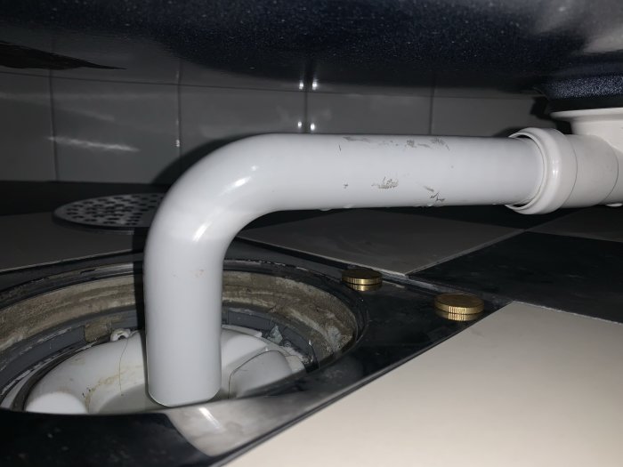 Närbild av en felinstallerad golvbrunn med synlig PVC-rörböj och nivåskillnader vid klinkern.
