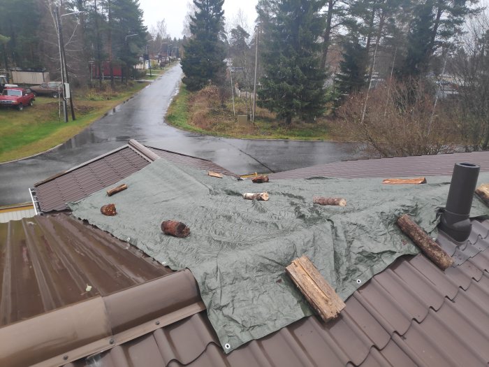 En presenning täcker en del av ett vått tak med stockar som tyngder, nära ett takfönster och avloppsventilation.