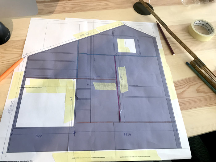 Arkitektskiss av en platsbyggd bokhylla med mått och markeringar på ett skrivbord med måttband och pennor.