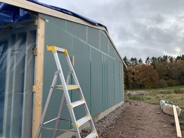 Delvis färdigställd garagevägg med utegips och grön tejp längs skarvar, stege framför och öppen landskap i bakgrunden.