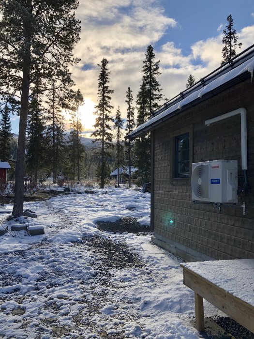 En nyinstallerad luftvärmepump på en husvägg med snötäckt mark och träd i bakgrunden under en solig dag.