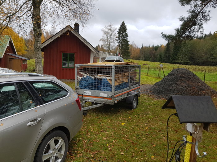 Släpvagn lastad med gipsplattor framför en silverfärgad bil, med en röd stuga och grushög i bakgrunden.