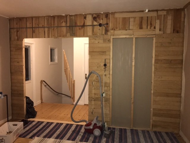 Inredningsbild av en trävägg med synliga takstolar och en öppning, en dammsugare framför, i ett hus från 1947.