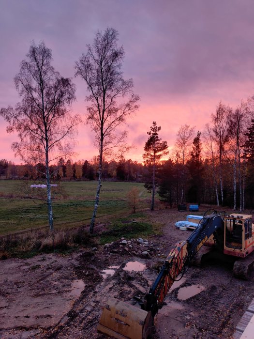 Dramatisk solnedgång med rosa himmel framför en byggarbetsplats med grävmaskin och båt i närheten.