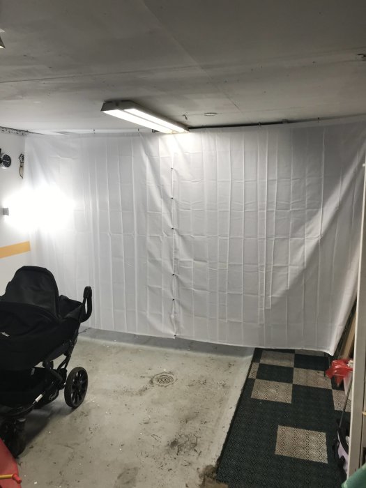 Vit plastduk uppsatt på vägg i garage med barnvagn och matta i förgrunden.