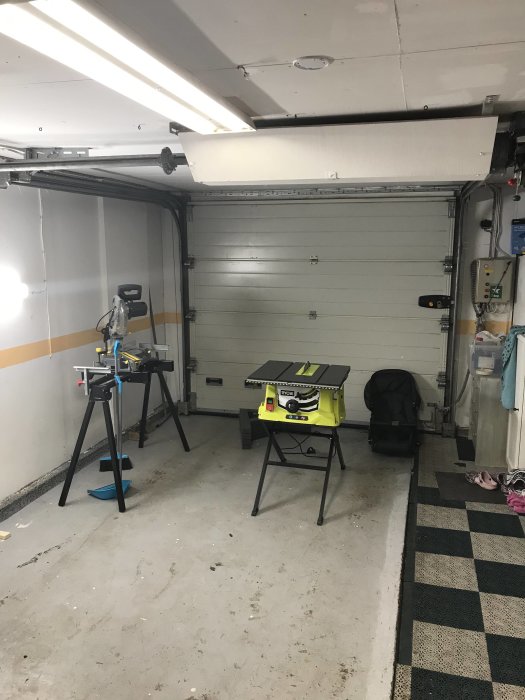 Garage med stängd port, arbetsbänkar, såg och olika verktyg, samt belysning i taket.