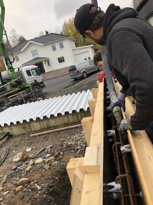 Arbetare fyller betong i träformar vid byggarbetsplats med betongbil och hus i bakgrunden.