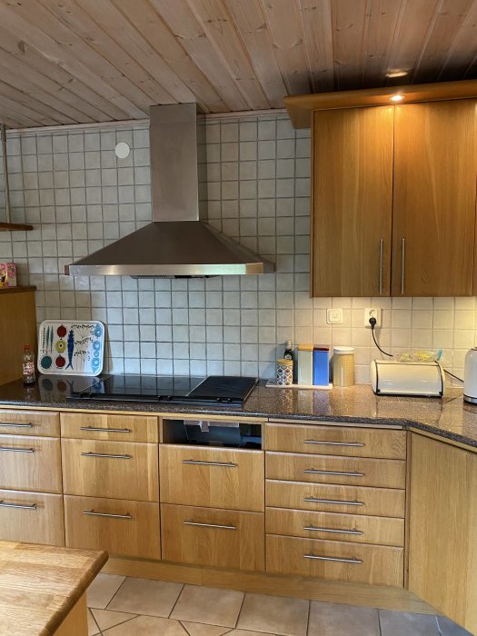 Ett kök med träskåp, rostfri spishäll, fläktkåpa och kaklade väggar, troligen från omkring år 2000.