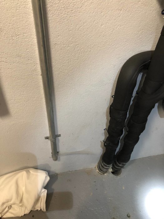 Metallrör för kablar och svart isolerat rör längs en källarvägg med synlig betongplatta.