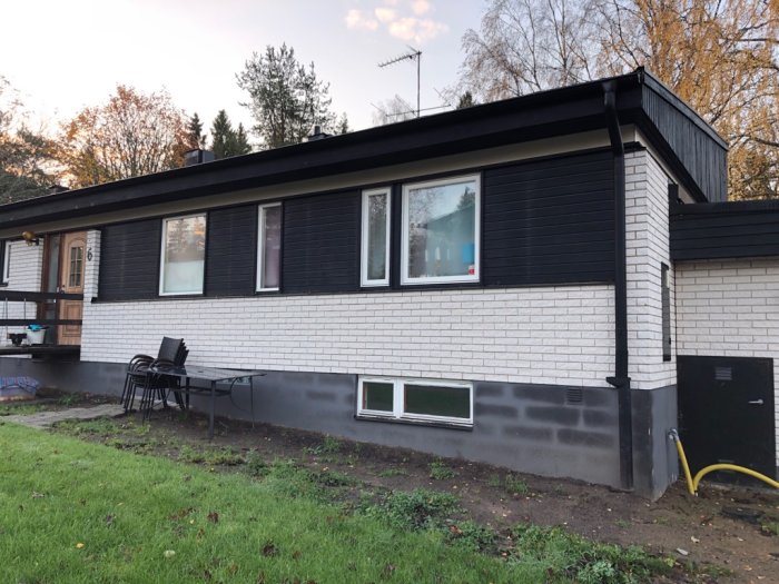 Renoverat enfamiljshus med ny fasad och svarta fönsterkarmar, uteplats med bord och stolar på gräsmatta.