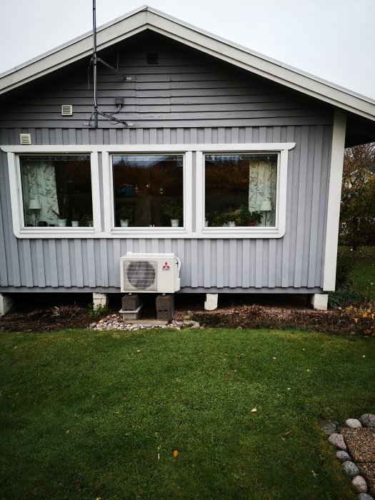 Framsidan av ett hus med blågrå panel och stora fönster, inklusive en värmepump placerad utanför.