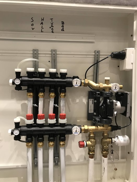 Värmesystemets fördelningsrör med termostater och etiketter för sovrum och badrum.