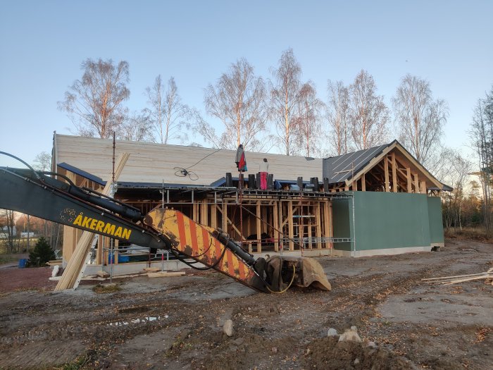 Byggplats med halvfärdigt hus där takarbeten precis är avslutade, med grävmaskin i förgrunden och klar himmel.