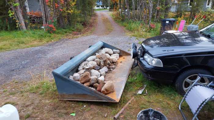 En släpvagn fylld med sten och grus framför en svart personbil på en grusväg, omgiven av grönska och trädgårdsredskap.