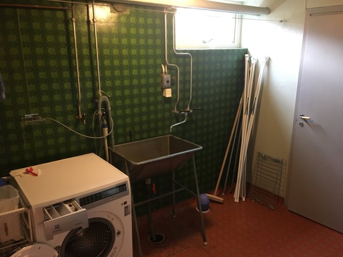 Gammaldags tvättstuga med grönt mönstrat tapet, röd matta, tvättmaskin, grovho och oinstallerade skåp.