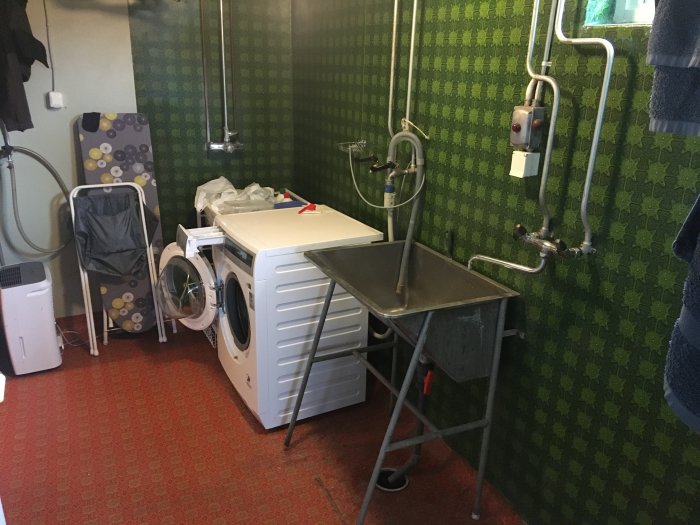 Tvättstuga med grön väggmatta, tvättmaskin, torktumlare och enkel diskbänk, behov av renovering.