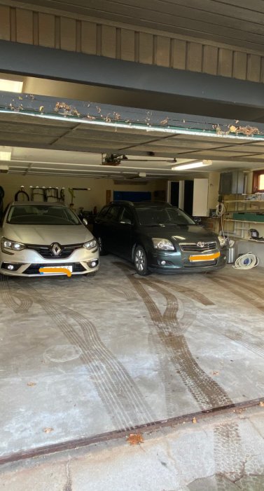 Två bilar parkerade i ett öppet garage med uppfälld port och verktyg på väggen.