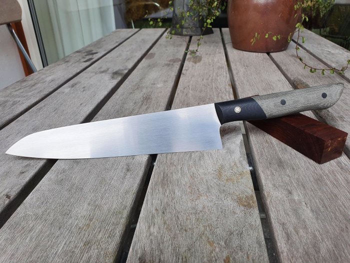 Handgjord kockkniv med 220 mm blad i guyto-stil på ett träbord med växt i bakgrunden.