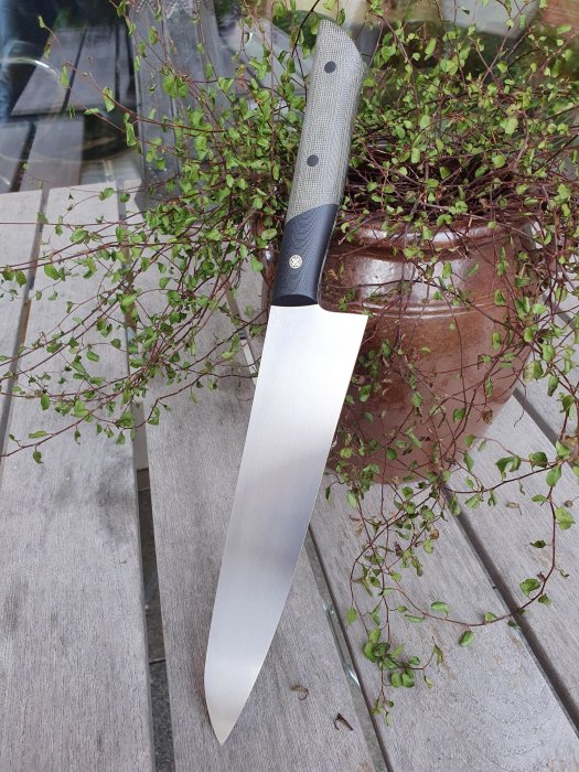 Handgjord kockkniv med 220 mm långt blad, placerad mot träbänk med en krukväxt i bakgrunden.