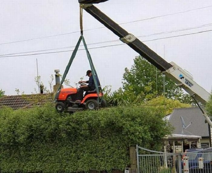 Person som sitter på en gräsklippare som lyfts av en kran över en häck.