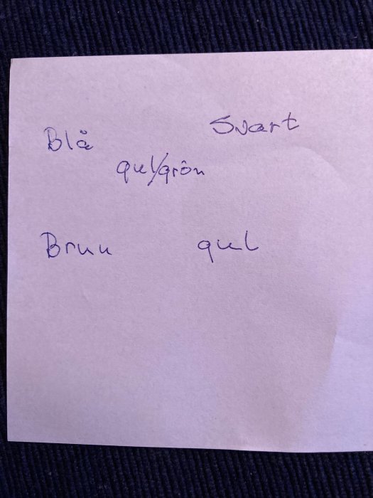 Handskrivna färger "blå, svart, brun, gul/grön" på ett vitt papper mot blå bakgrund.