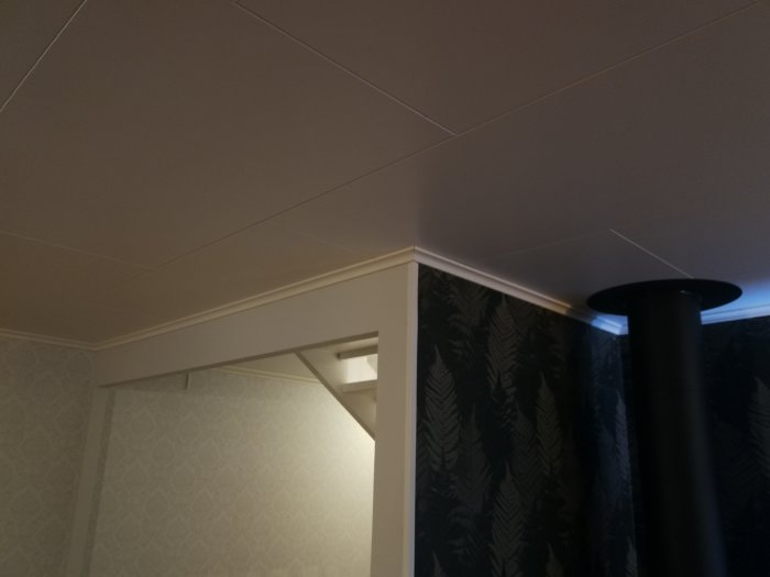 Innertak med synliga träbjälkar i en korsning, nära en svart pelare och tapetserade väggar.