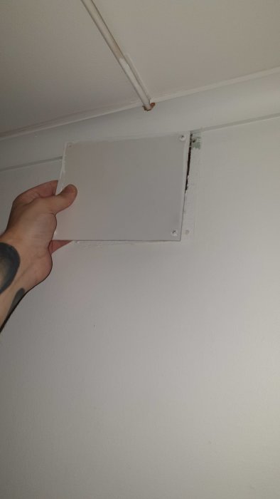 Hand öppnar en vit inspektionslucka i ett gipstak nära en elektrisk kabel.