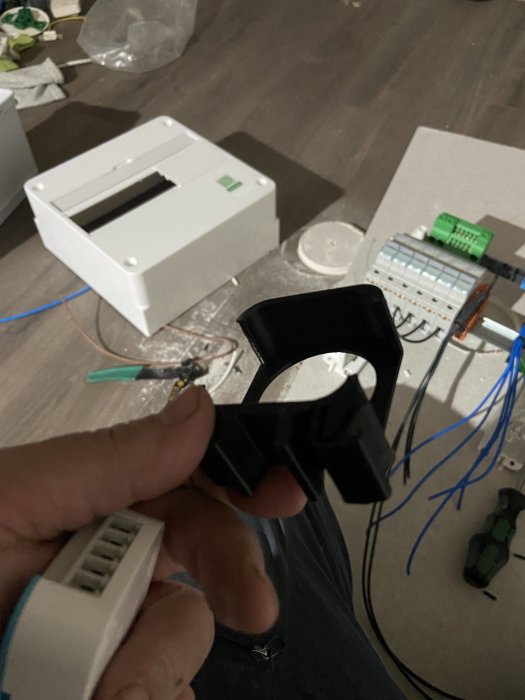 Hand håller upp svart 3D-utskrivet objekt med elektriska komponenter och kablar i bakgrunden.