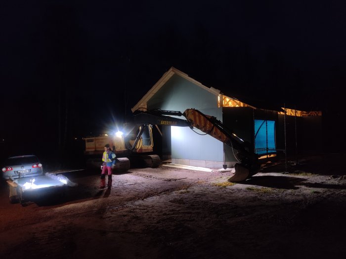 Grävmaskin med lyft arm som används som lyftkran för att flytta byggmaterial vid husbygge på kvällen.