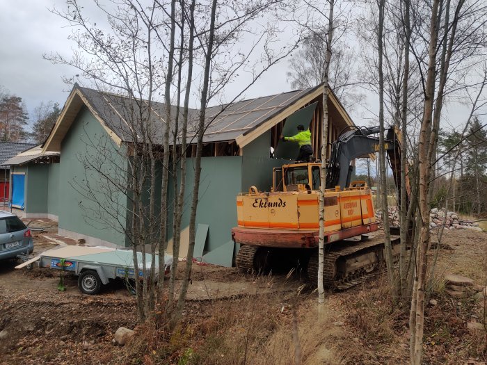 Hus under konstruktion med en grävmaskin vid gaveln där personer arbetar med byggställningar och gipsplattor.