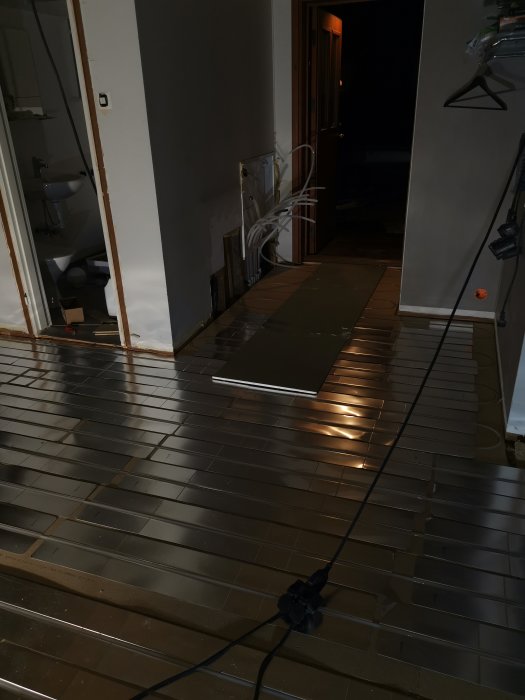 Golvvärmeinstallation med synliga slingor under mörka klinkerplattor i en korridor, sladdar och verktyg synliga.
