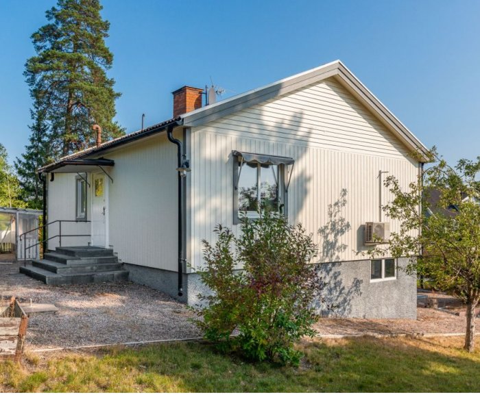 Ett vitt hus från 1945 med T11-tegel och platt entrétak mot en klarblå himmel, omgivet av grönska och en grusad framsida.