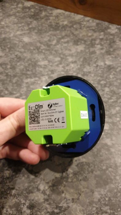 Hand håller en grön och blå ECOdim smart LED-dimmer på ett grått underlag.