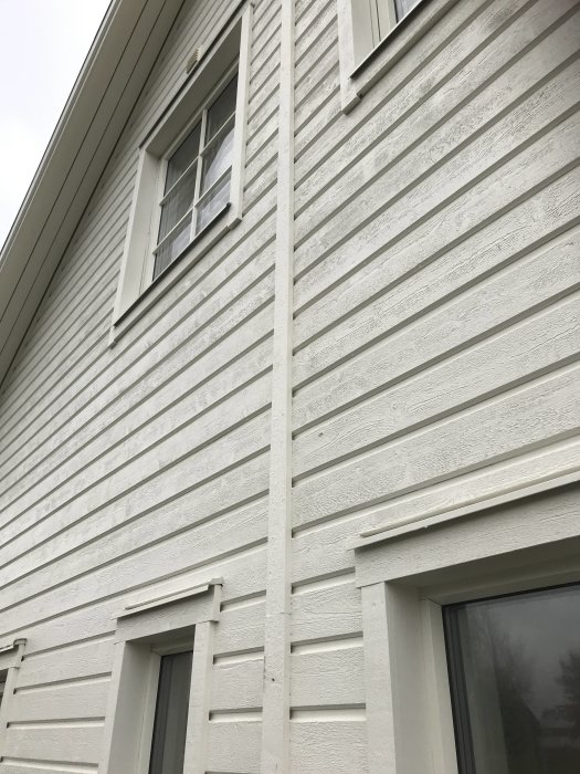 Vinkelvy av en vit husfasad med liggande panel och flera fönster, med fokus på anslutningen till taket.