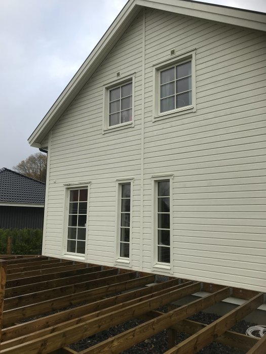 Husfasad med liggande panel och fönster, oskyddat träregelverk för uterum i förgrunden.