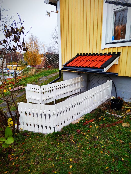 Nymålat vitt staket med dekorativa spjälor vid källarentren, omgivet av gräsmatta och en gul husvägg.