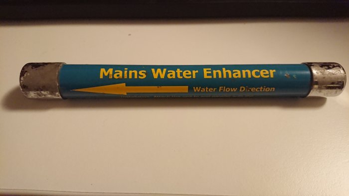 Avlägsnat hushålls vattenförbättrare med text "Mains Water Enhancer" och pil för vattenflödesriktning.