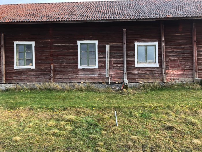 Gammal ladugårdsbyggnad med rödfärgade väggar och tre fönster, synlig stengrund som sjunkit och en lyft med domkraft.