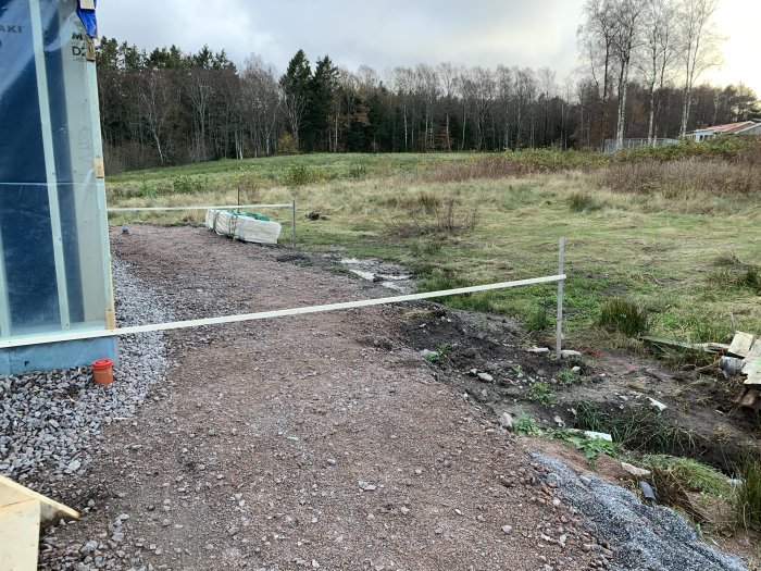 Plats förberedd för gjutning av plintar bredvid hus, markerad med snören, i ett ruralt landskap.