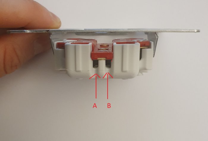 Hand håller en eluttag mot en vit bakgrund, två markerade anslutningspunkter A och B för jordning.