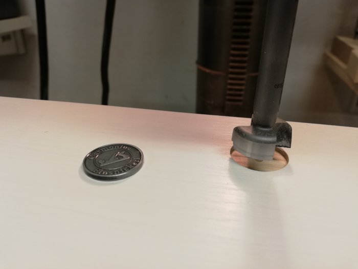 Egendesignat mynt intill ett 20mm hål infäst med epoxi i träyta, verktyg för montering.