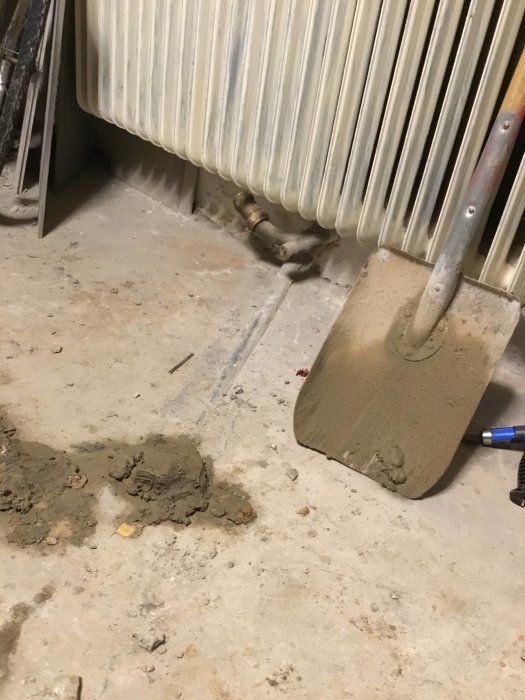 En spade lutad mot en radiator, cement på golvet som indikerar pågående arbete med rörledningen.