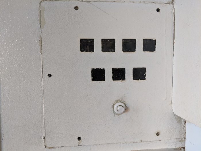 Gammal vit knapppanel med åtta svarta knappar och en ringklocka, viss lack och smuts.
