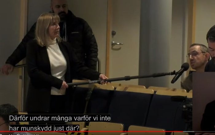 Person står vid podium i samtal om användning av munskydd i kollektivtrafiken, textning syns i förgrunden.