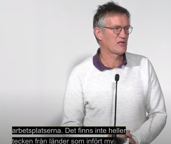 Man i mikrofon talar om munskydd och smittspridning, textat på svenska.