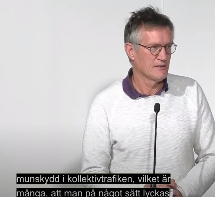 Man i vit tröja med mikrofon föreläser, texten på skärmen diskuterar munskydd i kollektivtrafik.