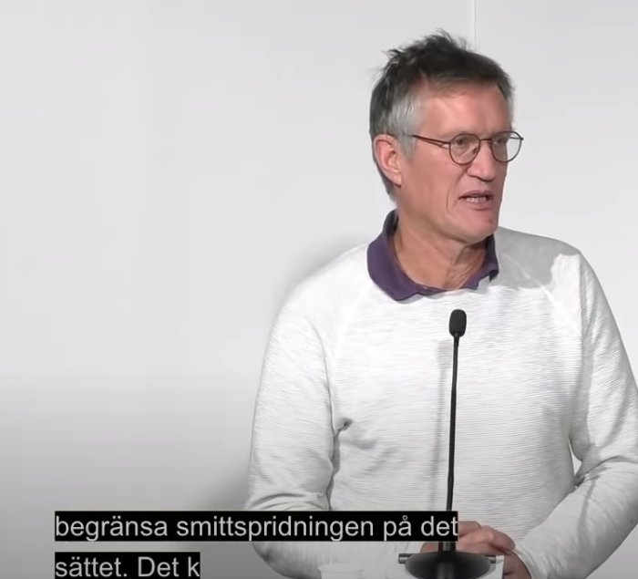 Man i vit tröja med mikrofon talar, svensk text om smittspridning synlig i bild.