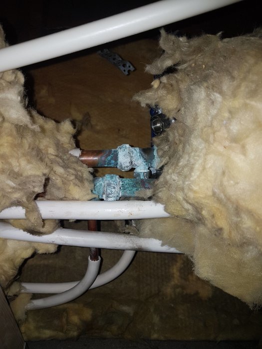 Korroderade rörkopplingar omgivna av isoleringsmaterial och vita plaströr.